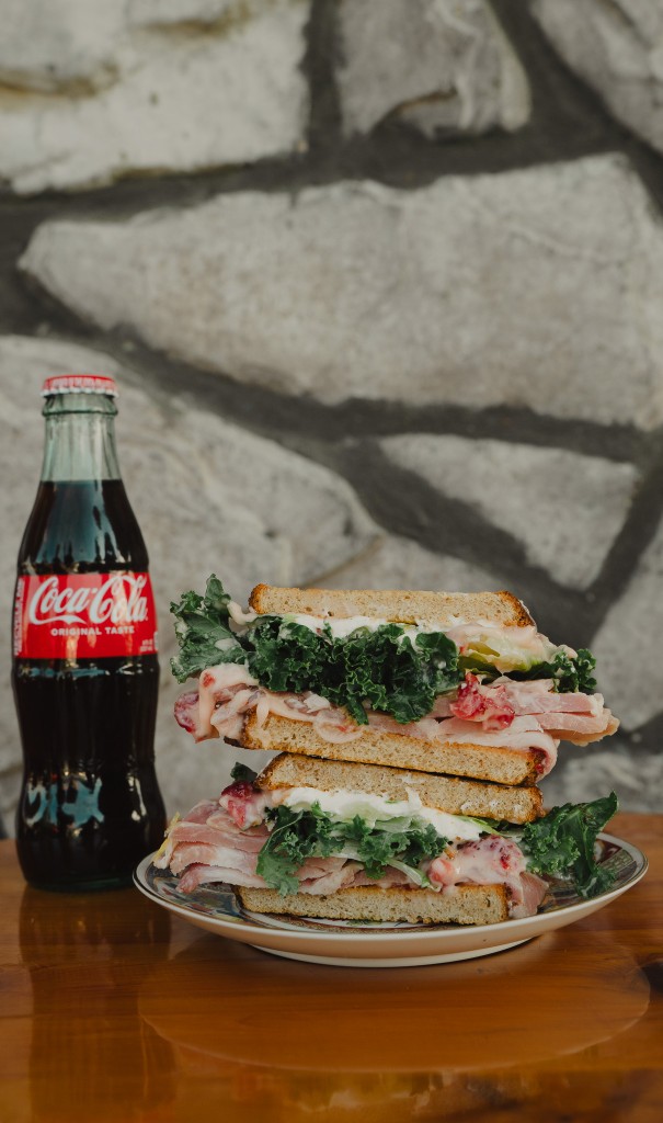 Stacked deli sandwich on a plate with a glass bottle of Coca-Cola, stone wall and wood table